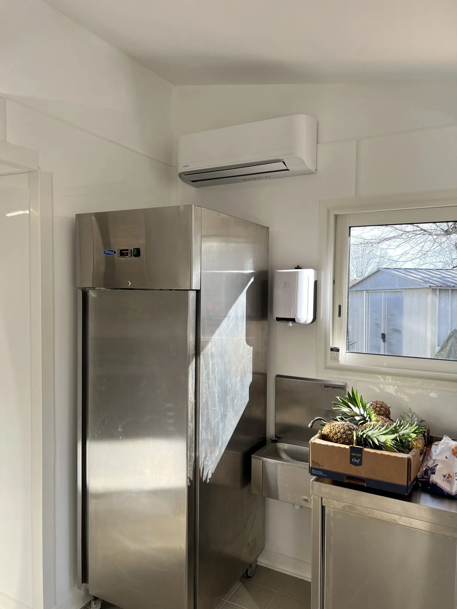 Petite cuisine professionnelle avec armoire réfrigérée inox, climatisation murale et carton d'ananas