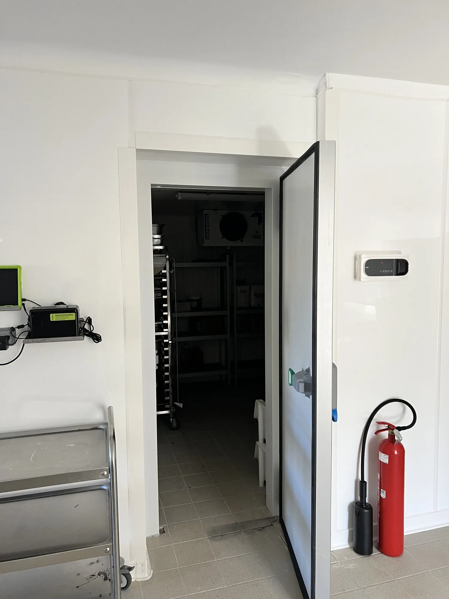 Porte isotherme pivotante blanche ouverte sur le local de stockage carrelé, extincteur et chariot inox