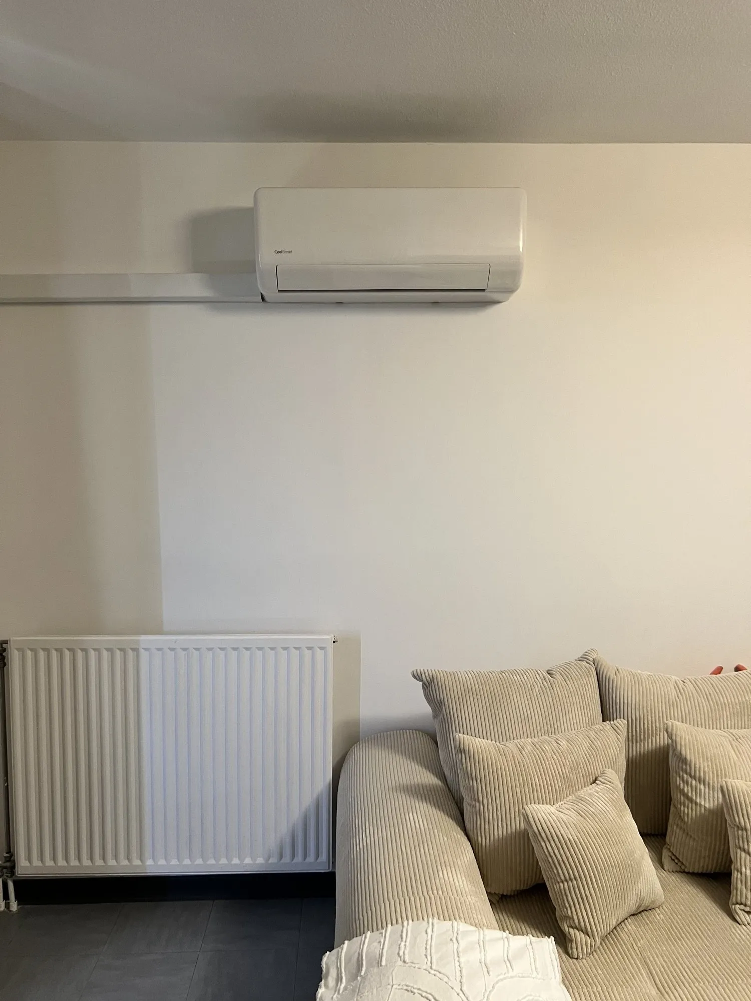 Unité intérieure de climatisation murale blanche au-dessus d'un radiateur, dans un séjour au canapé beige