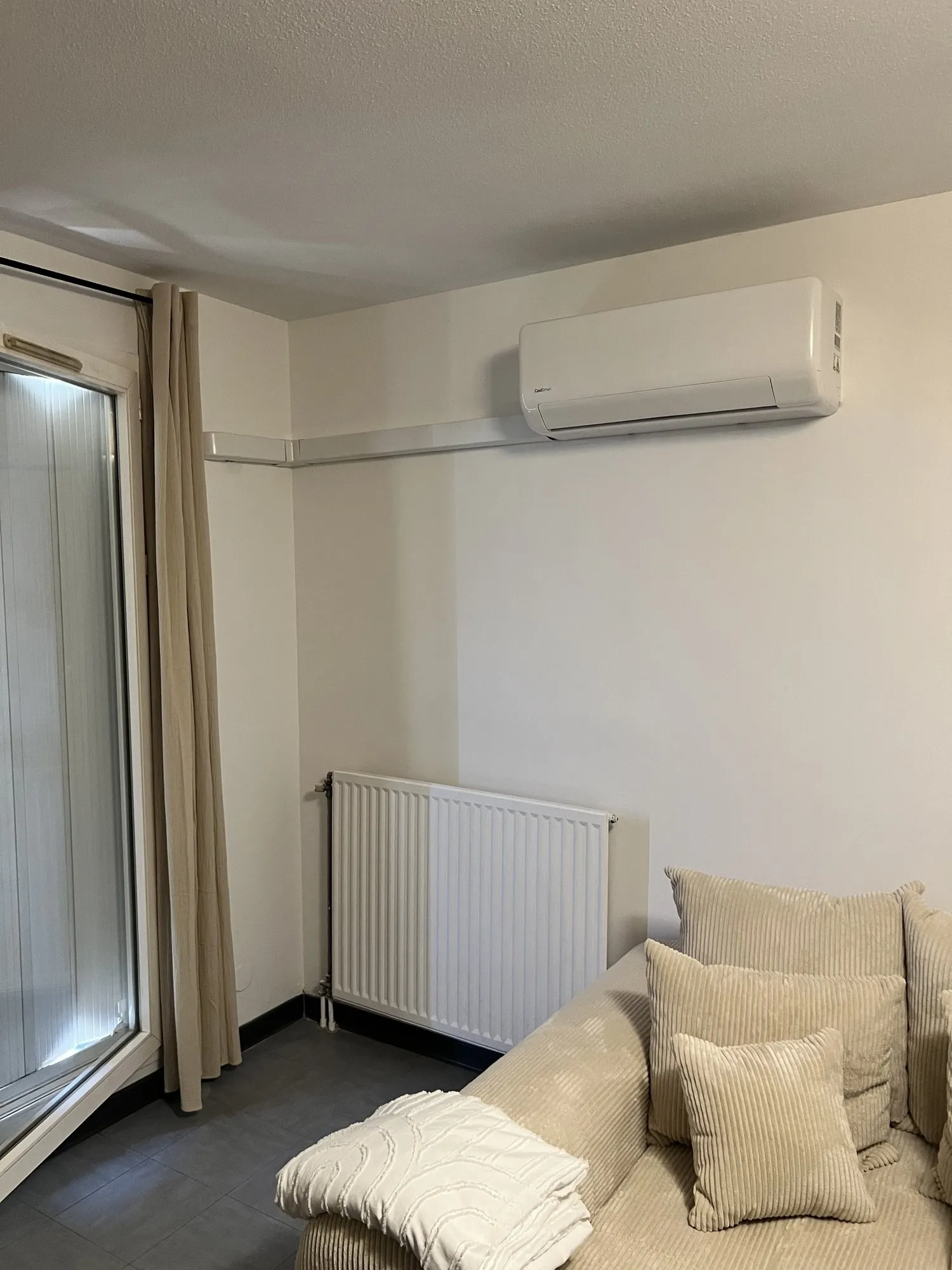 Unité murale de climatisation installée près d'une porte-fenêtre, radiateur et canapé en dessous