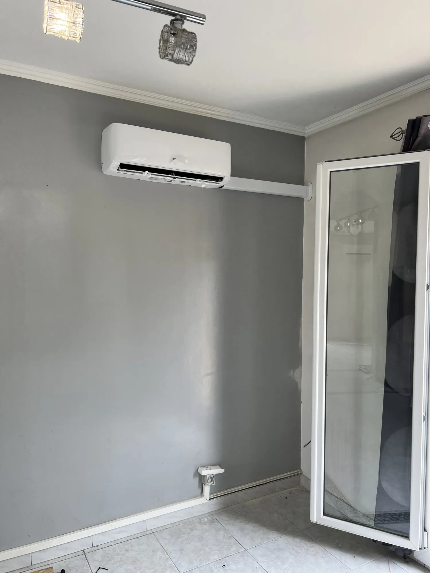 Unité intérieure de climatisation murale sur un mur gris, à côté d'une porte-fenêtre blanche