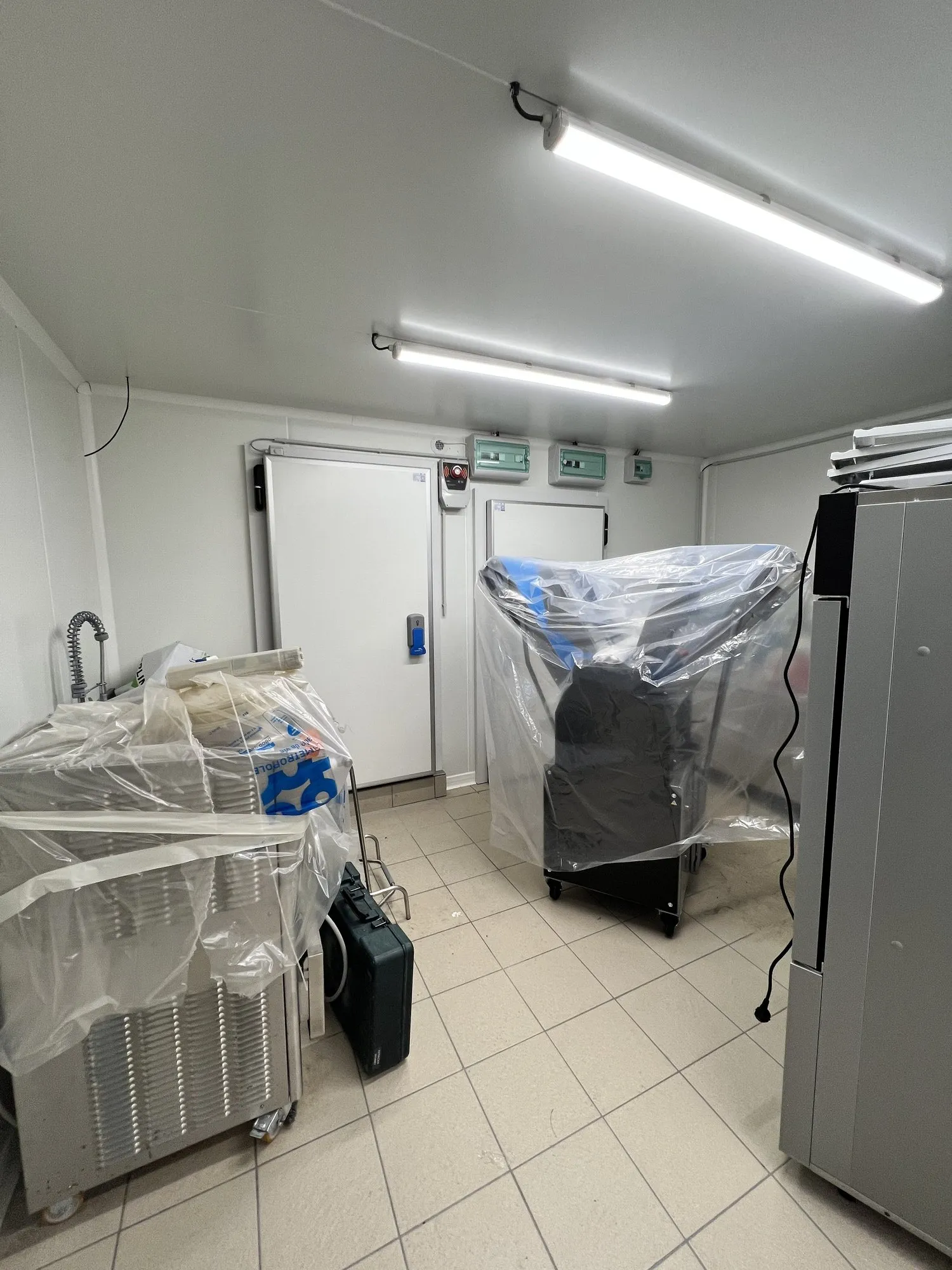 Porte de chambre froide blanche et équipements réfrigérés encore sous film plastique dans la cuisine carrelée