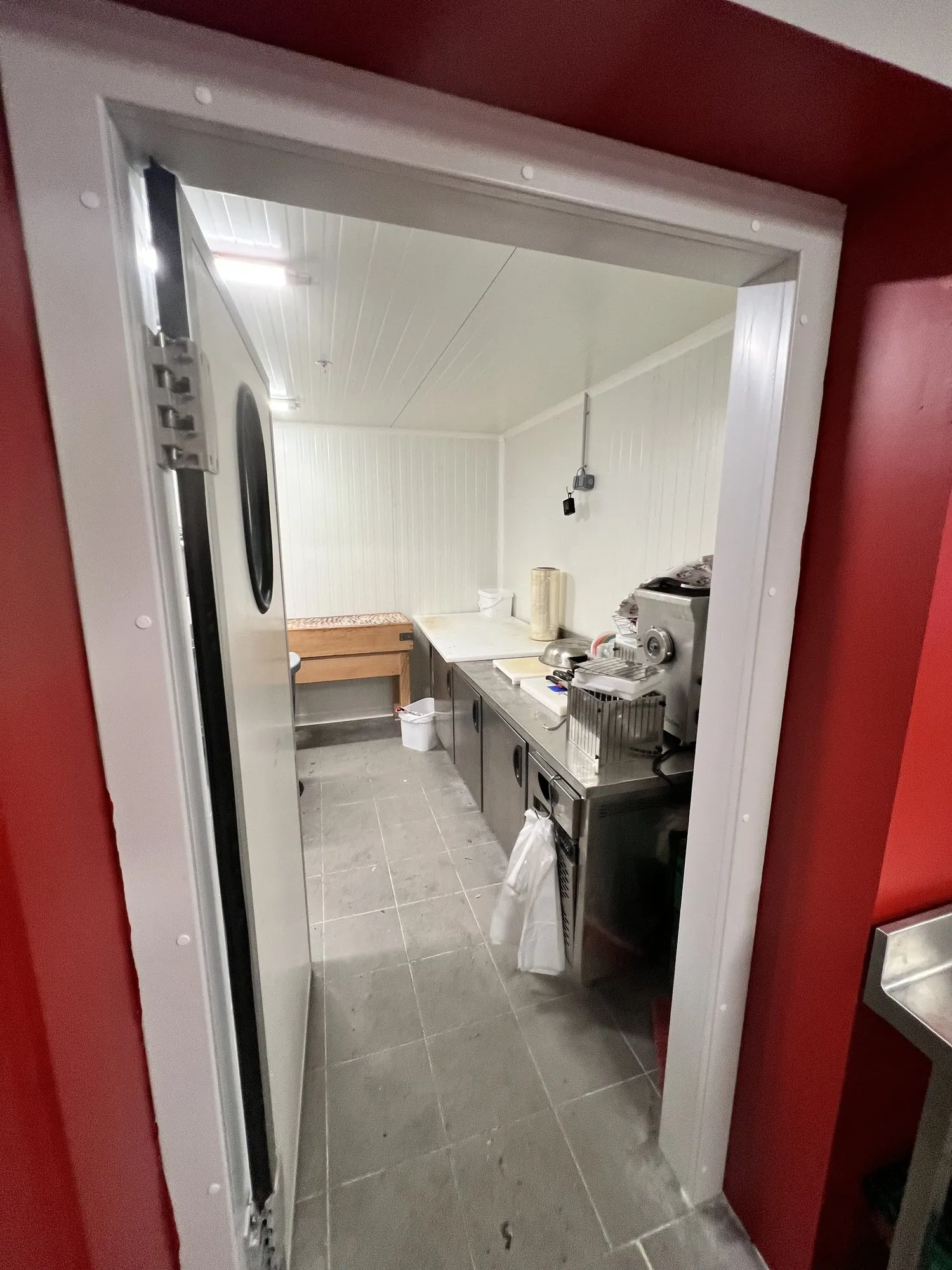 Vue depuis la porte isotherme vers la cuisine attenante : plan inox et appareils, encadrement rouge