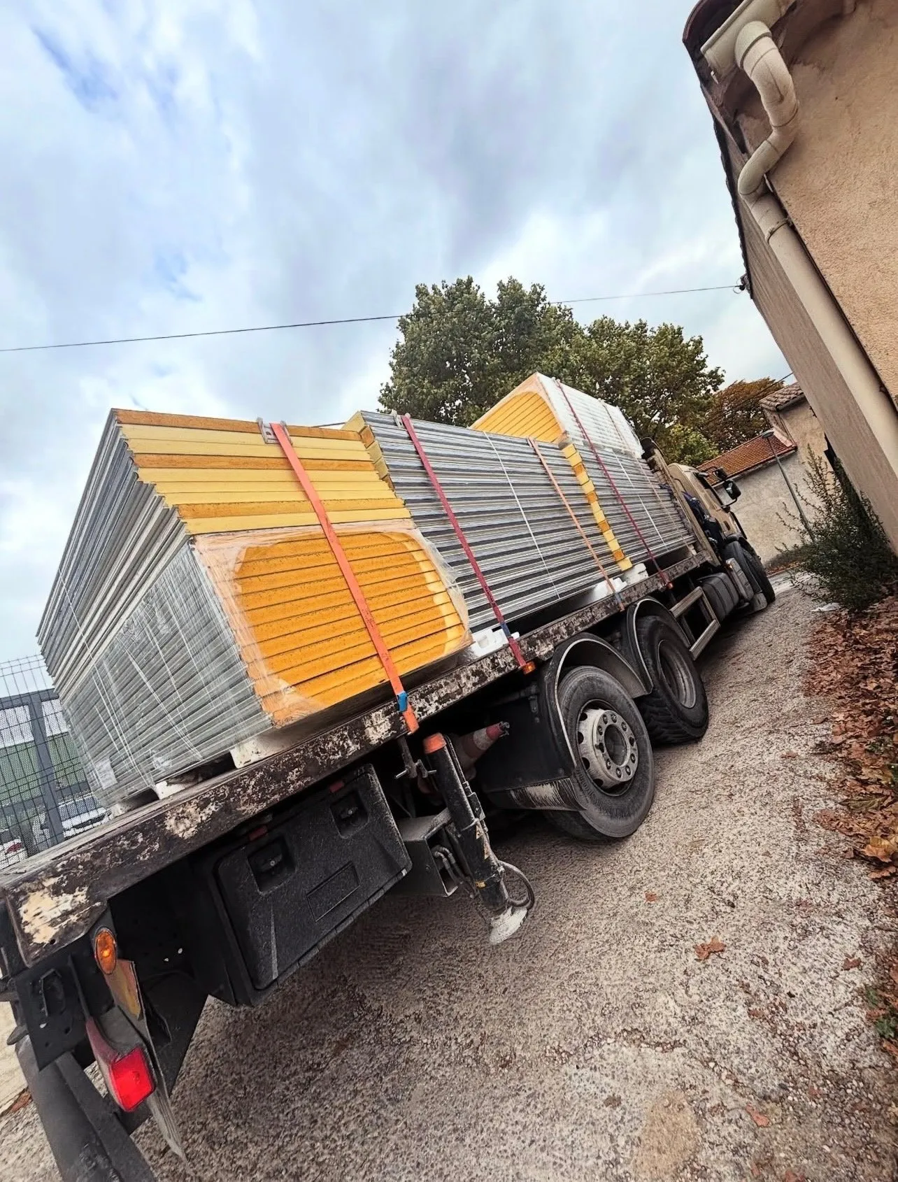 Camion plateau chargé de panneaux sandwich à âme jaune et de bardage métallique, sanglés, garé devant une maison