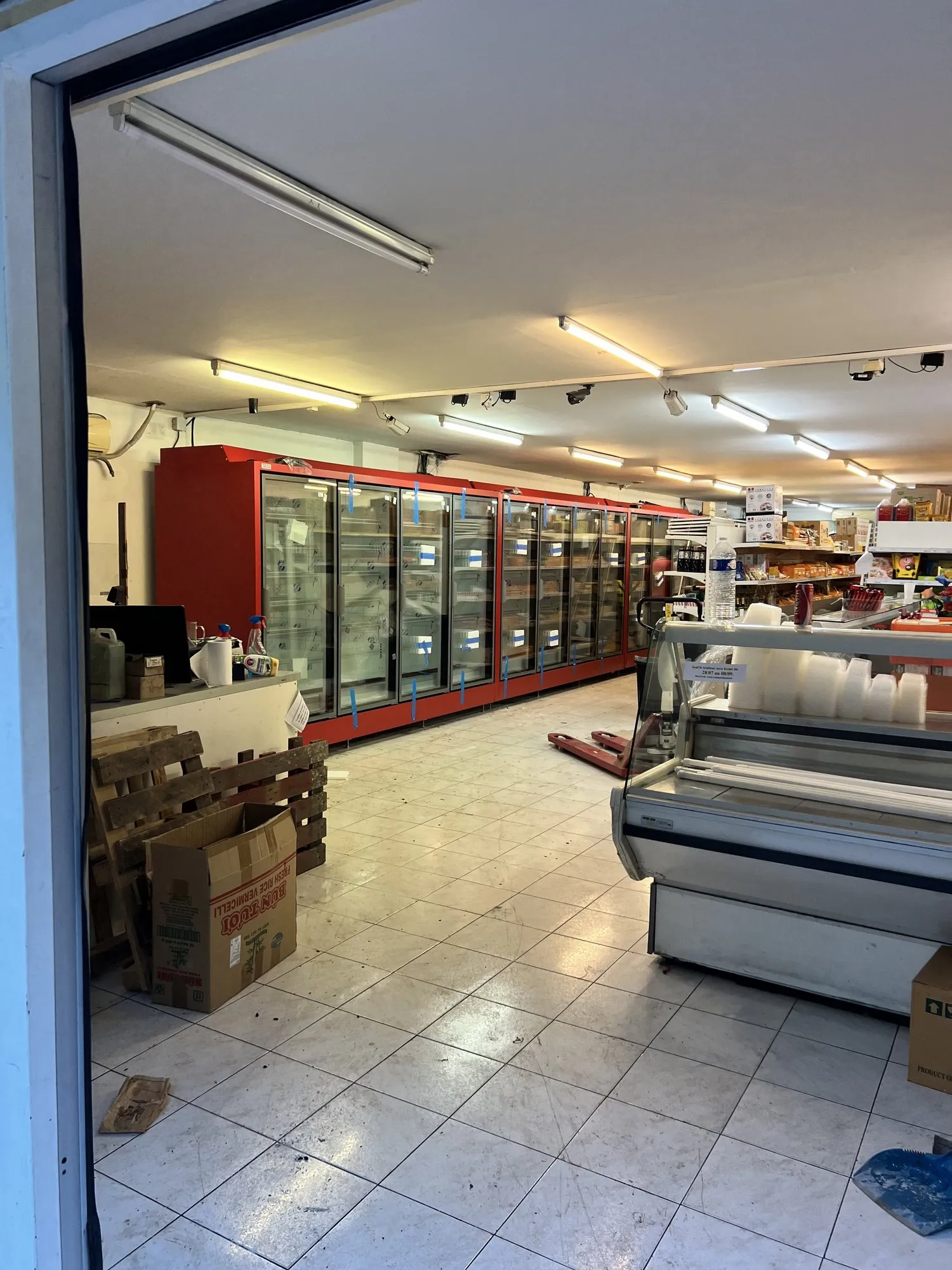 Épicerie vue depuis l'entrée : vitrines et armoires réfrigérées à cadres rouges le long du mur, cartons au sol pendant l'installation