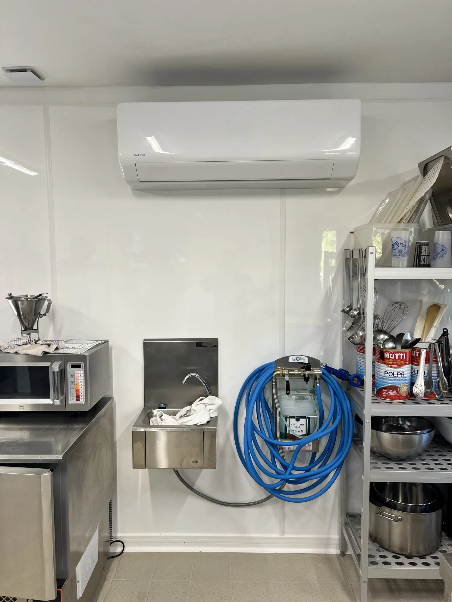 Cuisine professionnelle en inox : climatisation murale au-dessus d'un lave-mains, tuyau bleu et rayonnage