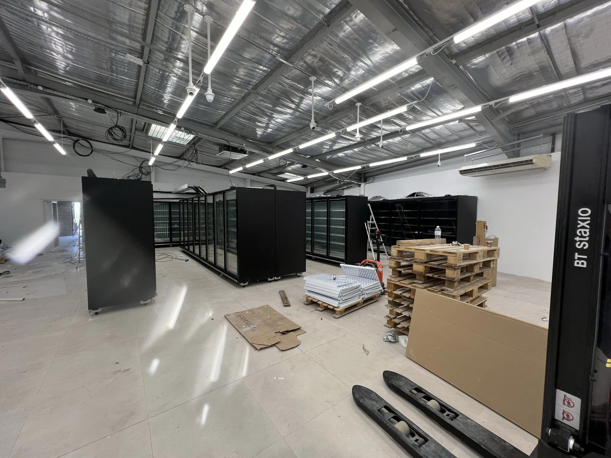 Magasin en travaux : meubles réfrigérés noirs en cours d'installation, palettes et outillage sous la charpente